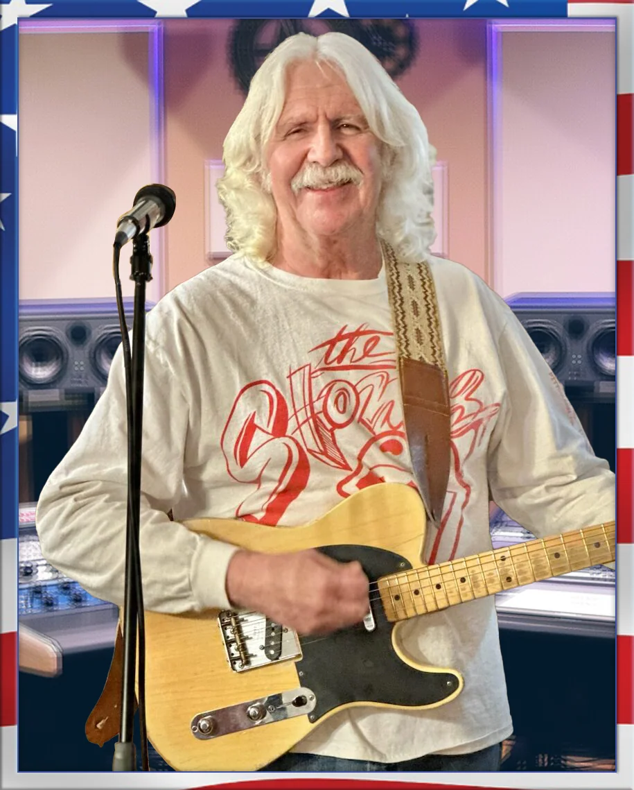 Man with white hair and mustache playing yellow electric guitar at microphone in studio setting with American flag border